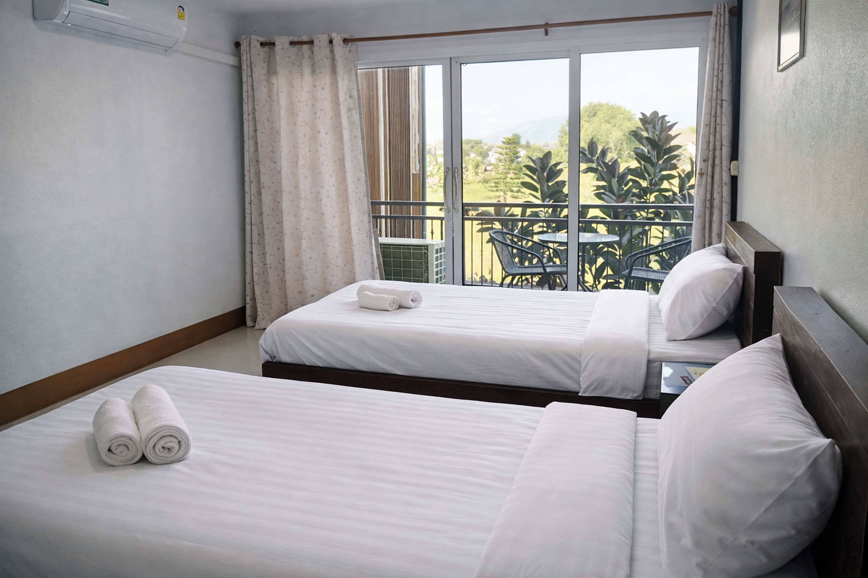 A hotel room with two beds, a balcony with a table and chairs, and a view of greenery and mountains.