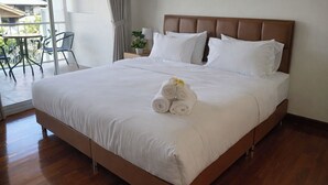 Un lit bien fait, avec du linge de lit blanc et des serviettes pliées, une tête de lit en bois et une vue sur le balcon, agrémenté de chaises et de plantes.