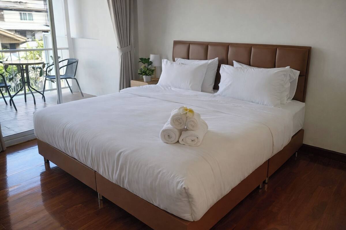 A neatly made bed with white linens and rolled towels, a wooden headboard, and a view of the balcony with chairs and plants.