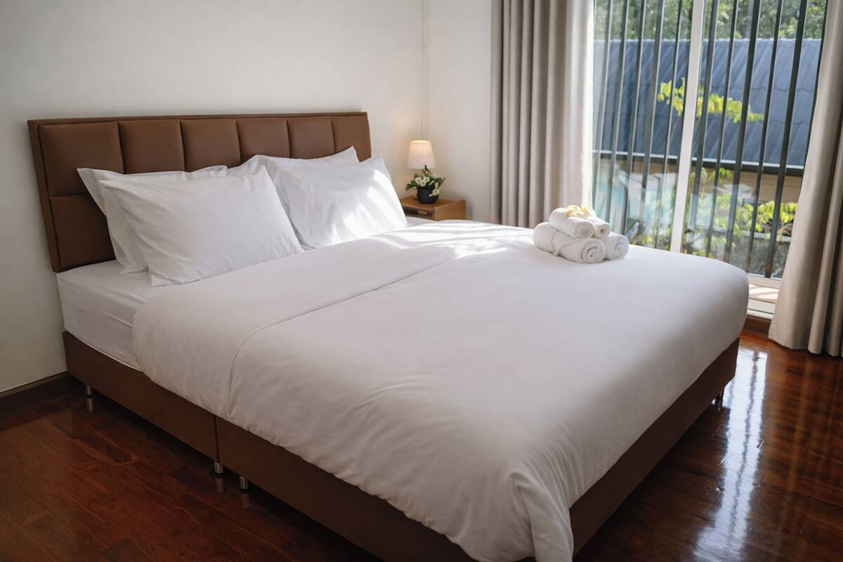 A neatly made bed with white linens and a brown headboard. There is a small bedside table with a lamp and a flower arrangement. The room has wooden flooring and a view of greenery outside through the window.