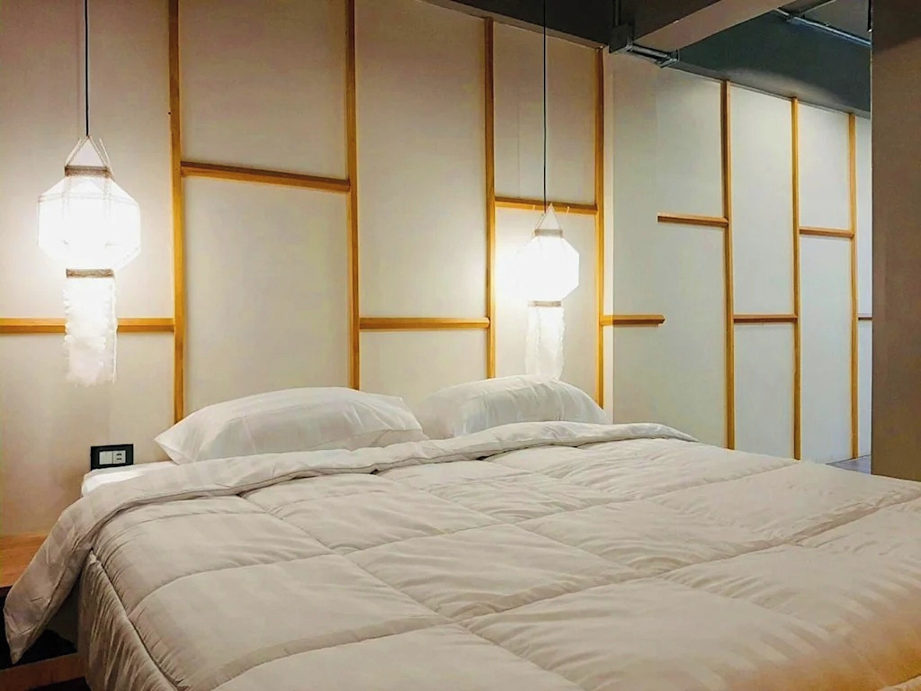 Un lit bien fait, avec une couette blanche, un lambris en bois sur le mur et deux lampes suspendues.