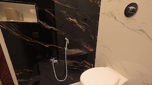 A bathroom with a white toilet, a shower with a handheld showerhead, and a marble wall. - Sasirekha Residency & Homestay (Puducherry)