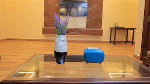 A glass-top table with a vase of purple flowers and a blue suitcase. - Sasirekha Residency & Homestay (Puducherry)