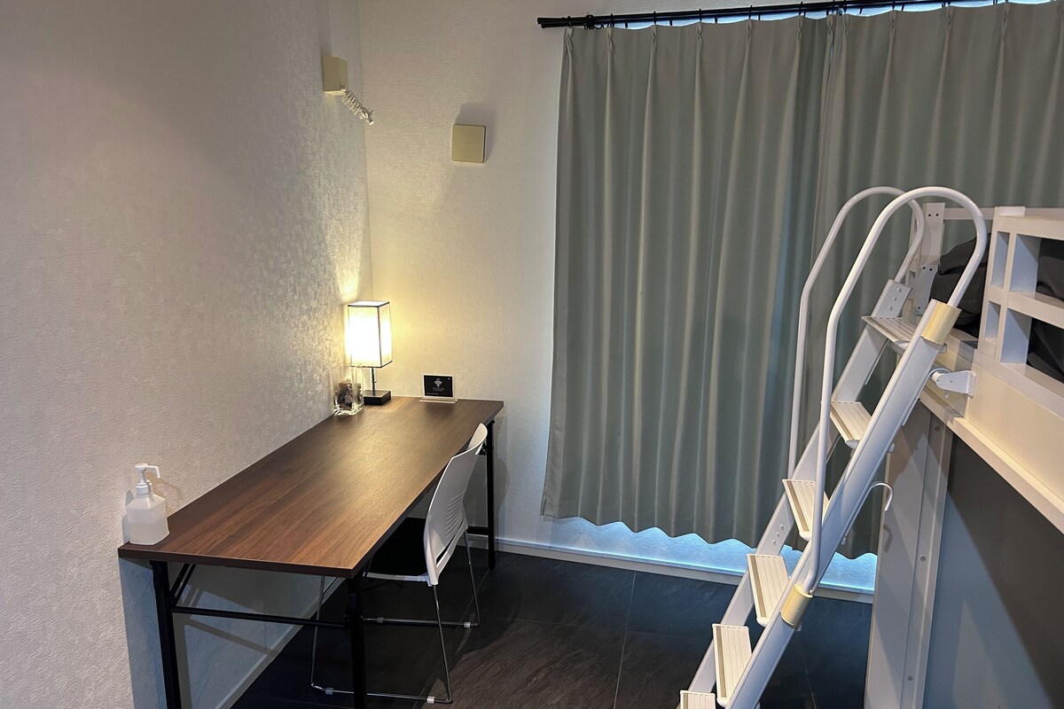 A room with a wooden table, a chair, a lamp, a small sign, and a window with curtains.
