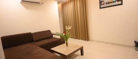 A living room with a brown sofa, a wooden coffee table with a vase of flowers, beige curtains, and a painting on the wall.