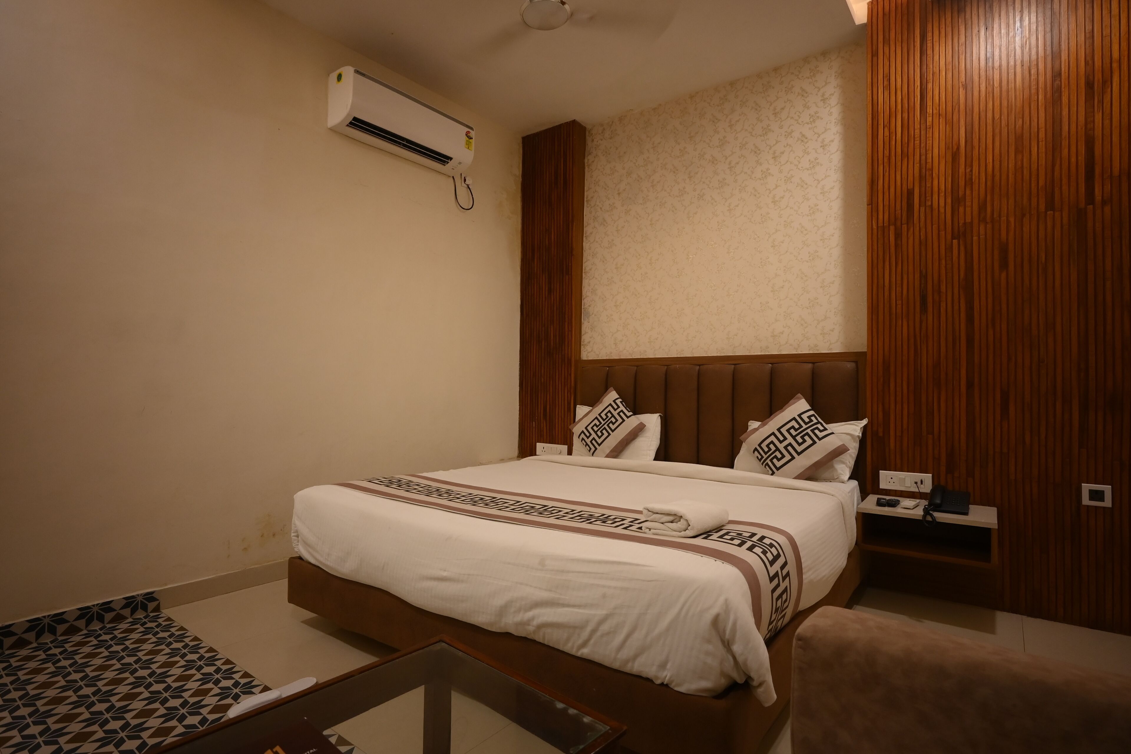A neatly made bed with a patterned bedspread, a bedside table with a phone and a remote, and an air conditioning unit mounted on the wall.