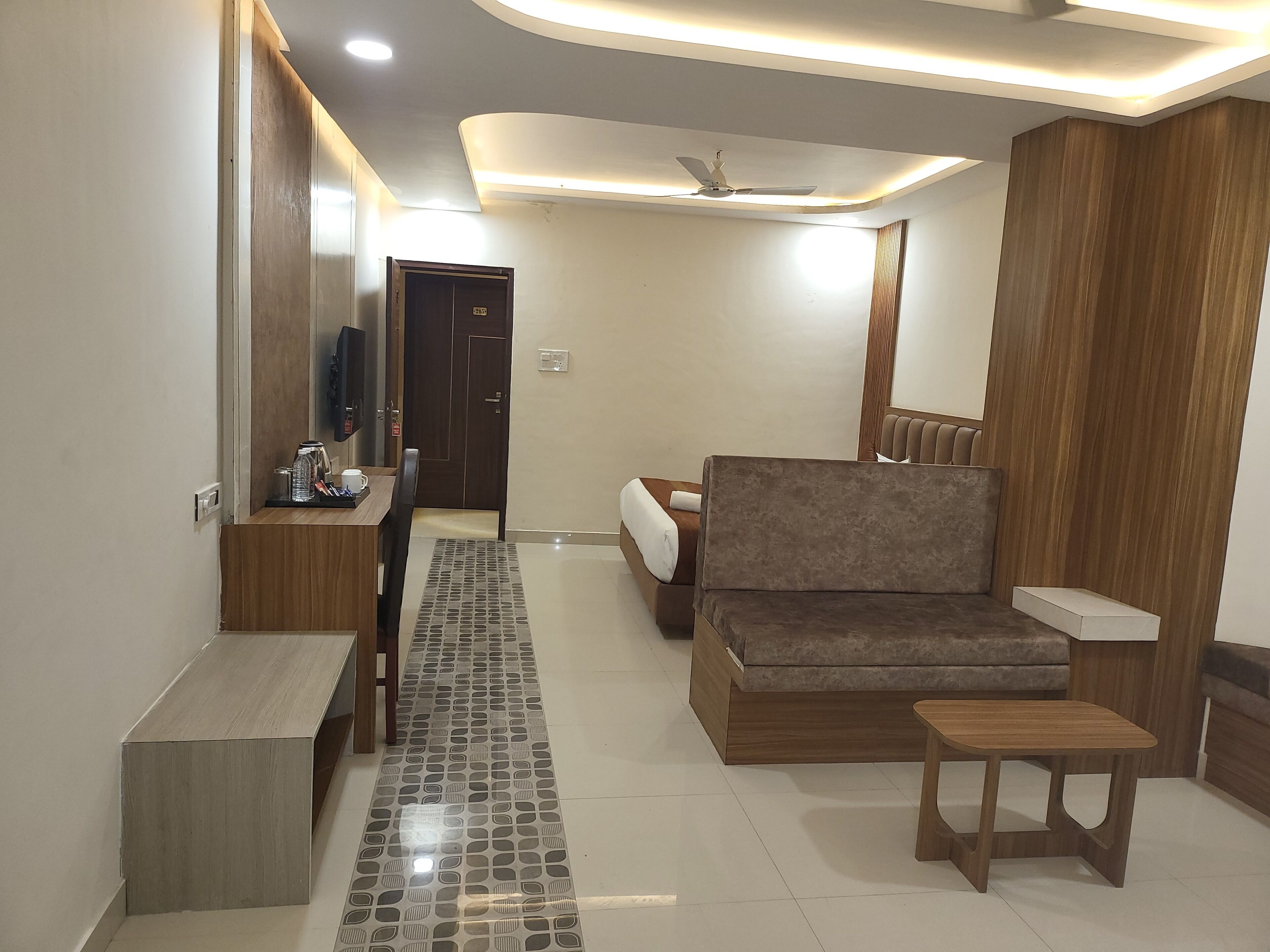 Habitación de hotel moderna con sofá, una mesita, televisor y baño con espejo y perchero para toallas.