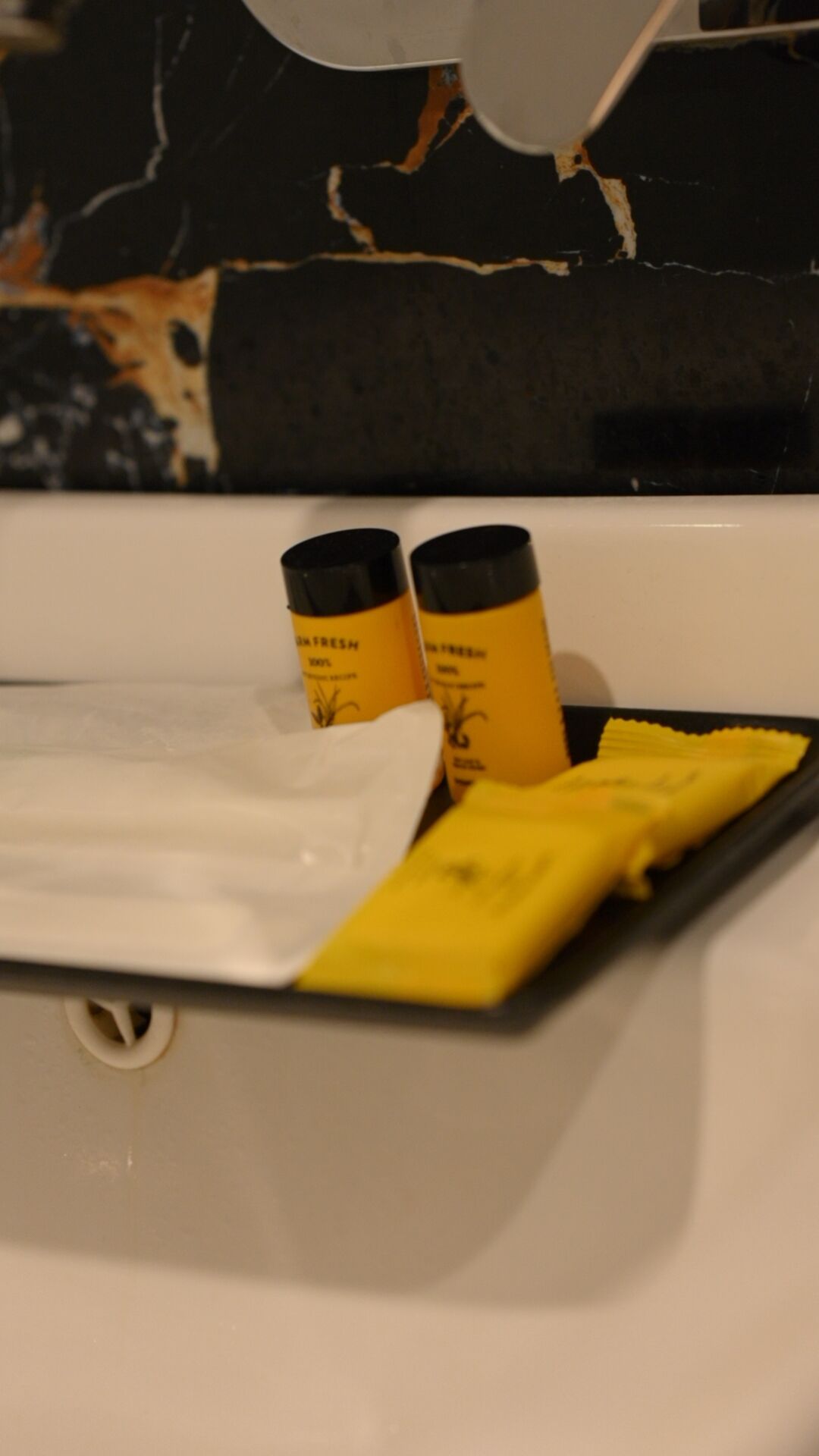 Ein Badezimmerwaschbecken mit einer weißen Spüle, zwei gelbe Flaschen mit dem Aufdruck "IN FRESH" und ein gelber Schwamm auf einer schwarzen Ablagefläche.