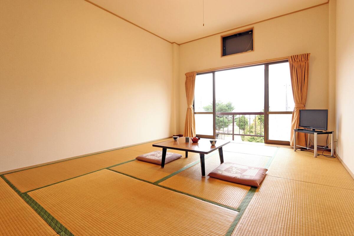 Een kamer met tatami vloerbedekking, een lage tafel met kussens, een televisie op een standaard en een raam met gordijnen.