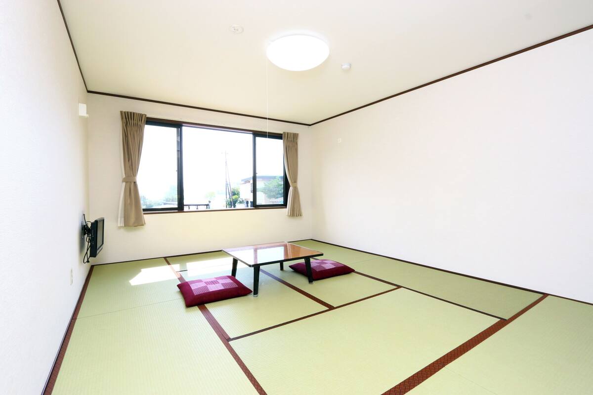 Een kamer met tatami vloer, een lage tafel, kussens, een raam met gordijnen en een plafondlamp.