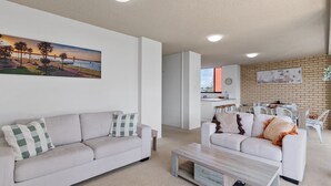 A modern living room with two sofas, a coffee table, and a kitchen area visible through an open door. - Tasman Towers Unit 8 (Port Macquarie)