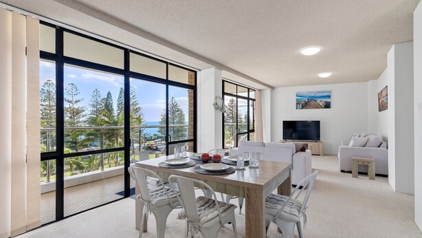 A modern dining area with a large table set for four, large windows offering a view of the ocean and trees, a flat-screen TV, and a cozy seating area with a sofa and armchairs. - Tasman Towers Unit 8 (Port Macquarie)