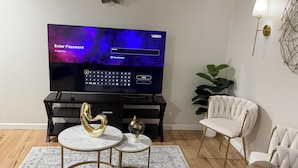 Una sala de estar moderna con un televisor de pantalla plana grande mostrando una pantalla de inicio, un sofá blanco, un sillón blanco, una pequeña mesa redonda con un adorno dorado y una alfombra estampada.