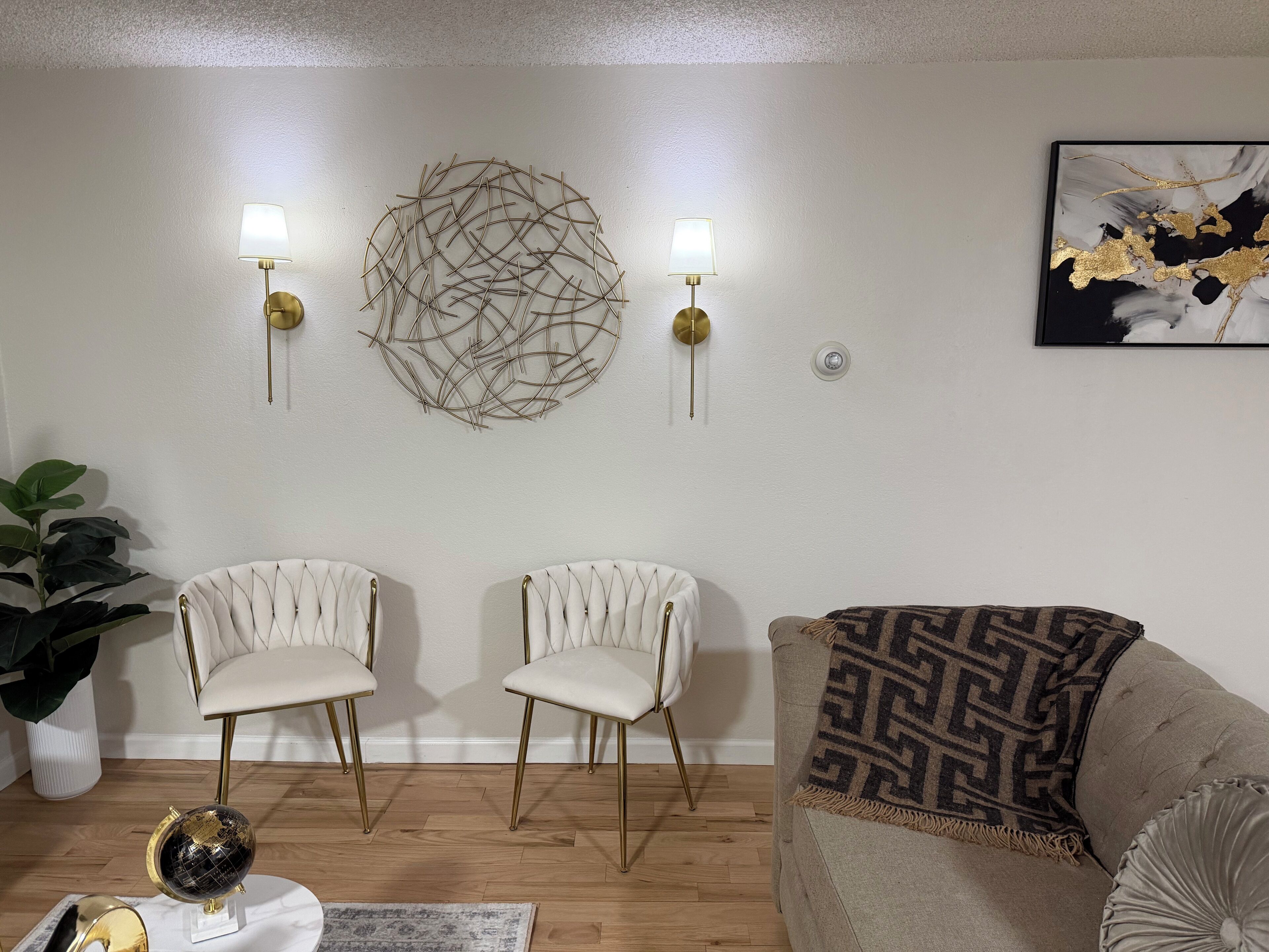 A living room with two white chairs with gold legs, a beige sofa with a patterned throw, a globe on a side table, and wall art.