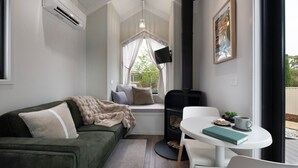 Een knus ingerichte kamer met een groene bank, een kleine tafel met een boek, een open haard en een raam met gordijnen.