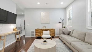 A modern living room with a wooden coffee table, a beige sofa, and a TV mounted on the wall. - Modern Apartment w/ Parking & Fast NYC PATH Access (Jersey City)