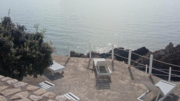 A coastal view with a rocky shoreline, a small island in the distance, and a seating area with benches and a railing. - Apartments Lux Lukić Utjeha (Utjeha)