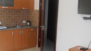 A kitchen with wooden cabinets, a sink, and a coffee maker. There is a door leading to another room and a television on the wall. - Apartments Lux Lukić Utjeha (Utjeha)
