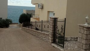 A coastal building with a stone wall and metal gate, a paved driveway, and a view of the sea. - Apartments Lux Lukić Utjeha (Utjeha)