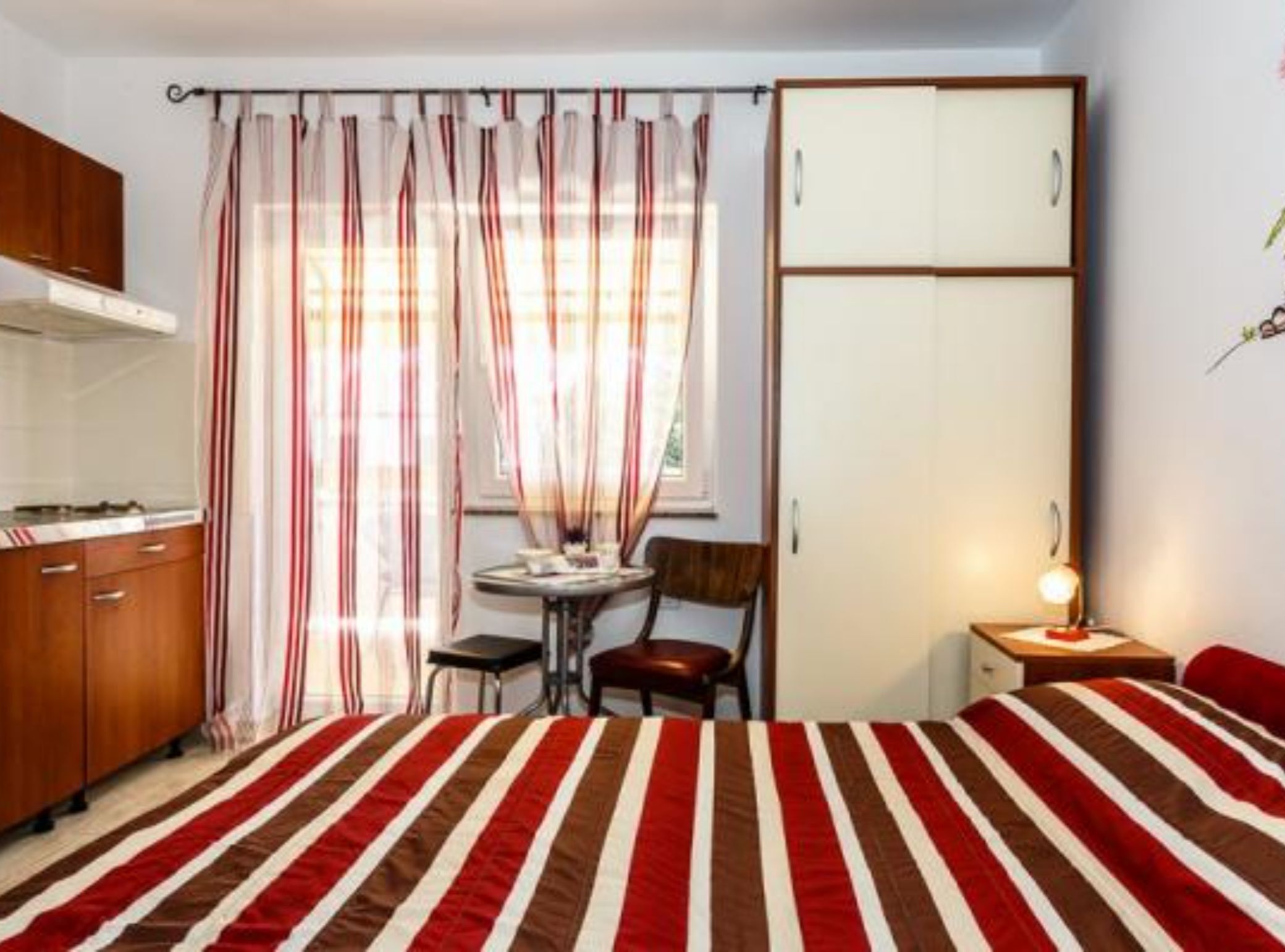 Quarto compacto com colcha listrada, guarda-roupa de madeira, mesa pequena com cadeira e abajur.