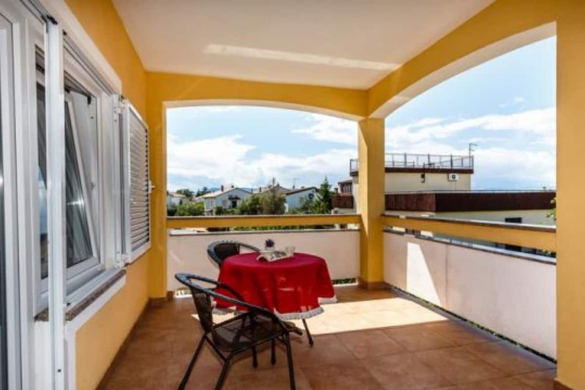 Un balcón con una mesa y sillas rojas, vista al cielo y edificios, y una pared amarilla.