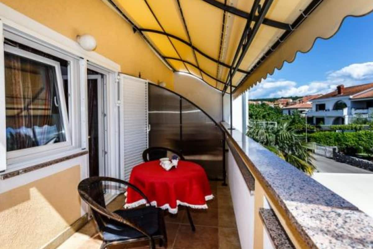 Un balcón con una mesa y sillas rojas, un toldo amarillo y vistas a edificios y vegetación.