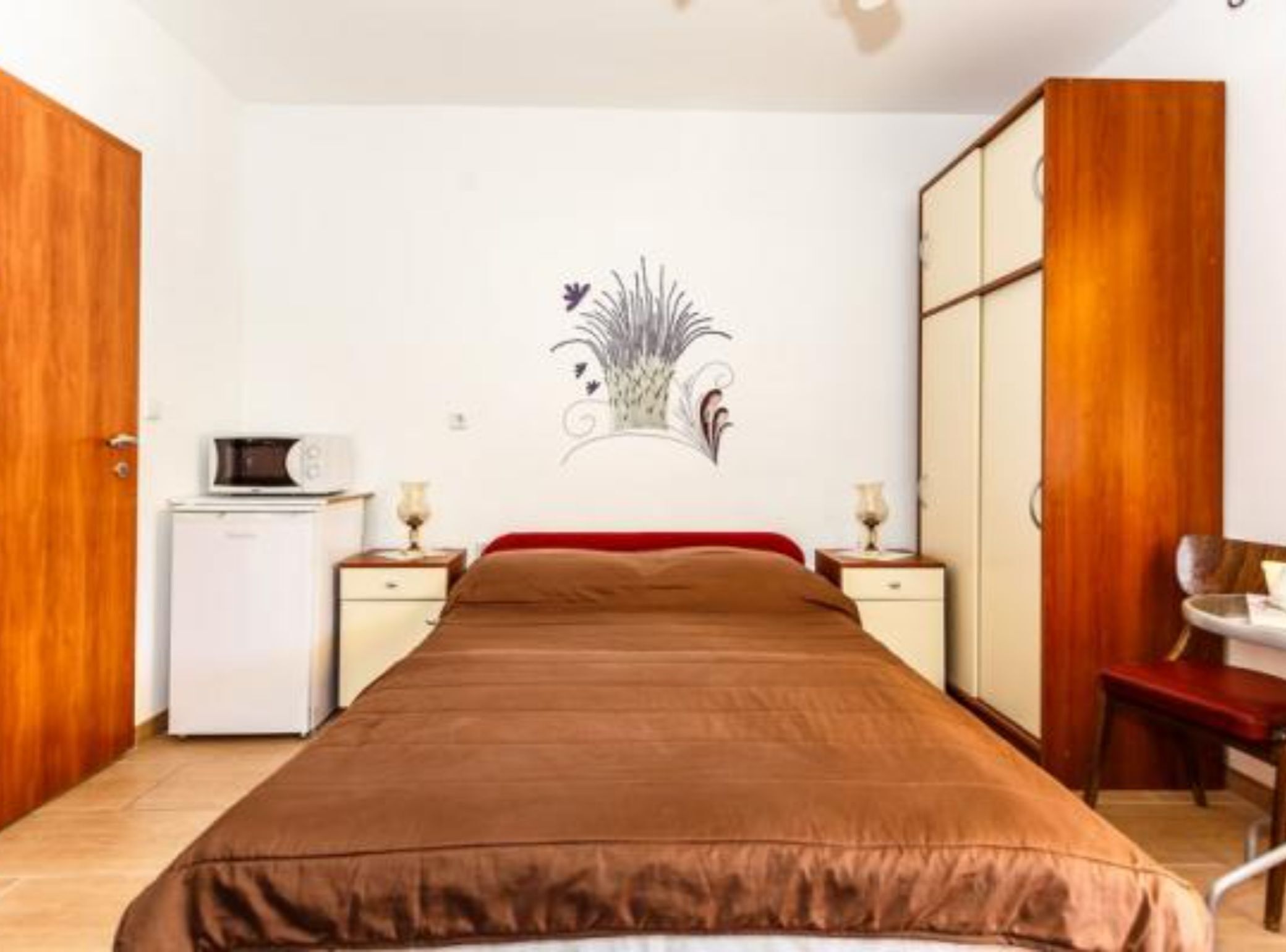 A bedroom with a bed, a desk, a chair, a wardrobe, and a small refrigerator.