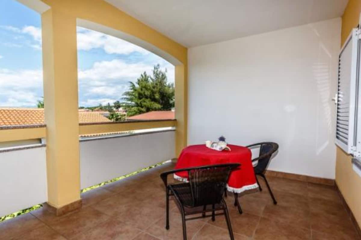 Un balcón con una mesa y sillas rojas, una ventana con persianas, y vistas al cielo y a los árboles.