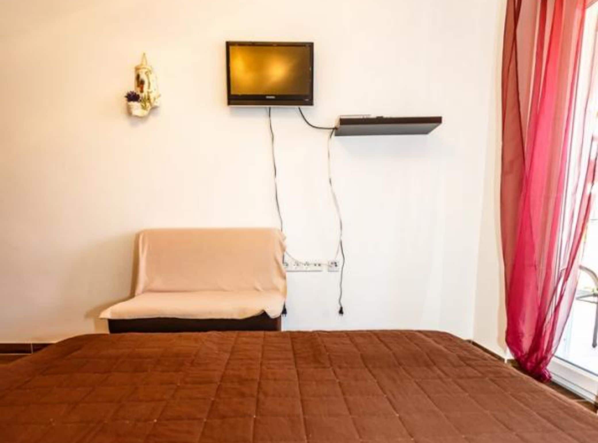 Ein Zimmer mit einem Bett, einem an der Wand montierten Fernseher, einem kleinen Regal und einem Fenster mit Vorhängen.