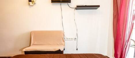 Ein Zimmer mit einem Bett, einem an der Wand montierten Fernseher, einem kleinen Regal und einem Fenster mit Vorhängen.