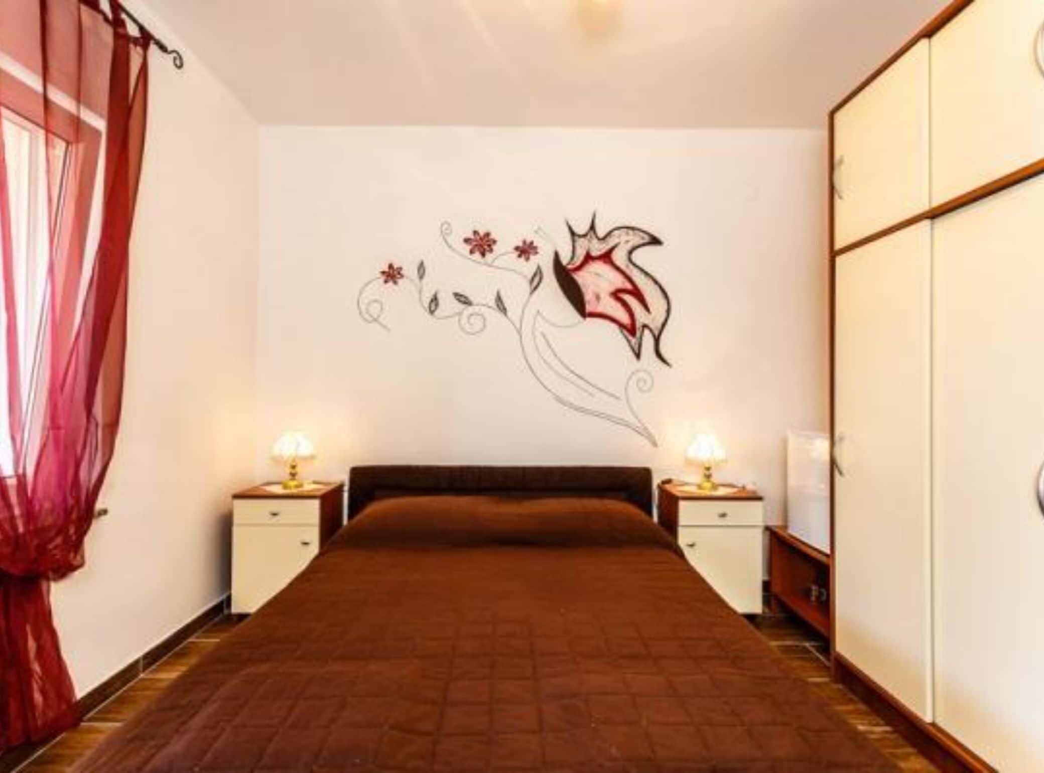 A bedroom with a bed, two bedside tables with lamps, a wall with a floral decal, and a wardrobe.