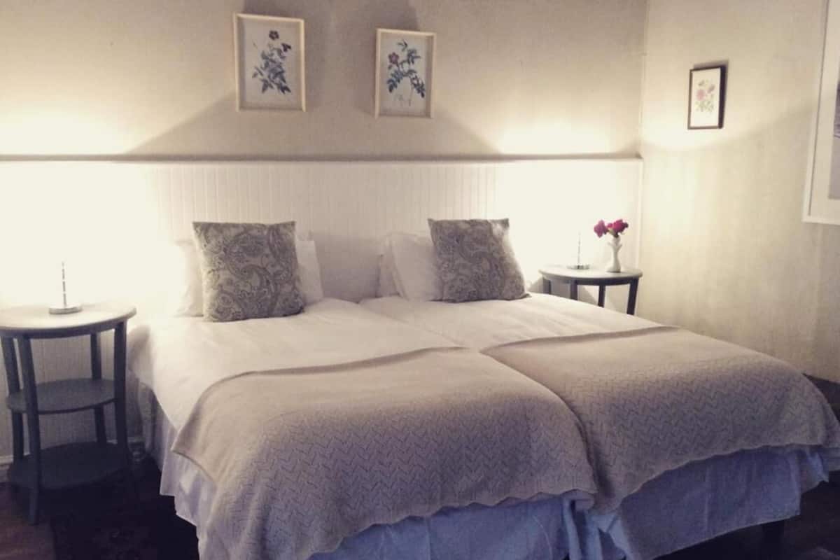 Una cama doble con una colcha blanca y dos almohadas, una mesita de noche con un florero y dos cuadros con motivos florales en la pared.