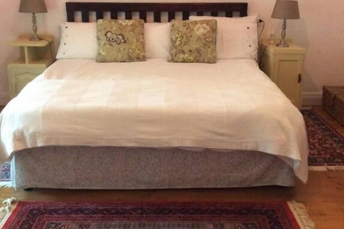 Una cama bien hecha con edredón blanco, dos almohadas a juego y un cabecero de madera. Hay dos mesitas de noche con lámparas, un tapete estampado y un espejo con marco decorativo fijado a la pared.