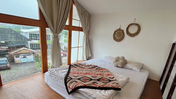 A bedroom with a large window, a bed with a patterned blanket, and two wall decorations. - Linggayoni Cottage Dieng (Dieng Kulon)