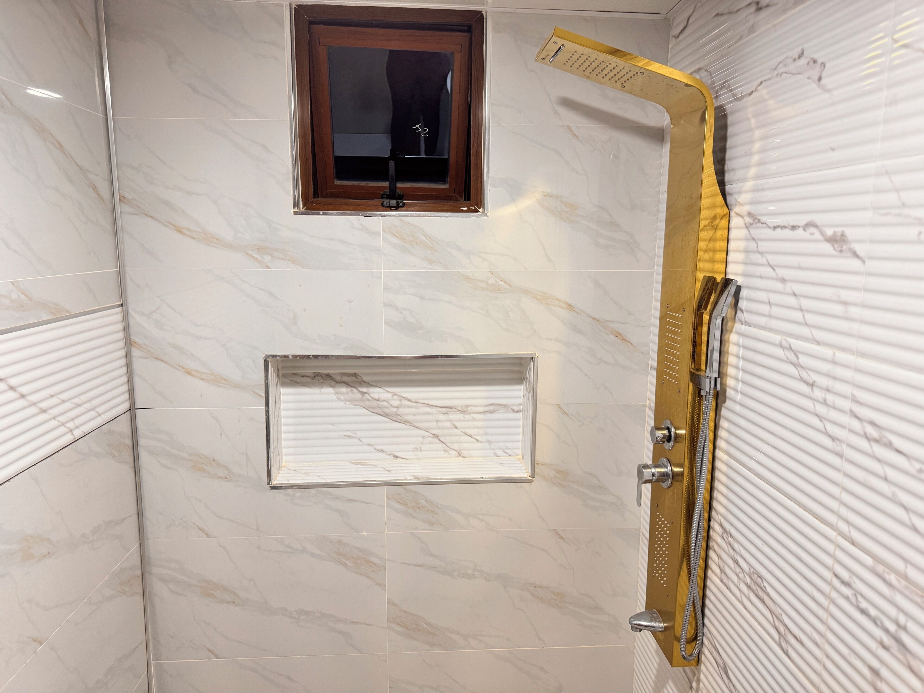 A bathroom with marble tiles, a window, a shower with a gold handle, and a towel rack.