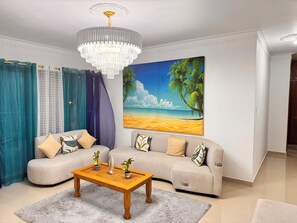 Een woonkamer met een beige bank, een houten salontafel, een kroonluchter en een schilderij met een strandthema.