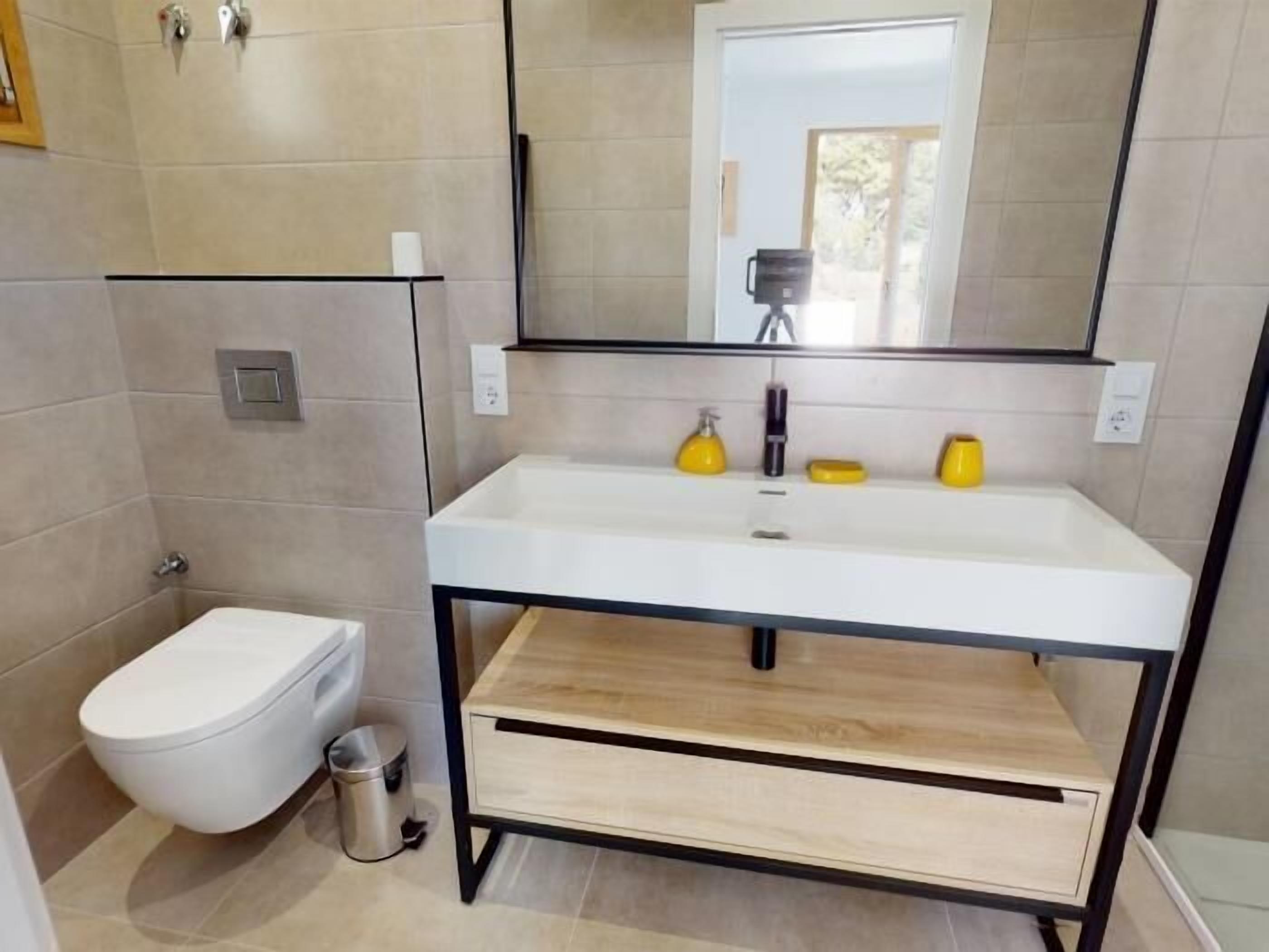Een moderne badkamer met een witte wastafel, gele zeepdispensers, een toilet en een spiegel.
