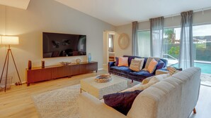 Una sala de estar moderna con un televisor de pantalla plana grande, un sofá azul oscuro con cojines estampados, un sofá beige, una mesa de centro de madera y un amplio ventanal con vista a una piscina.