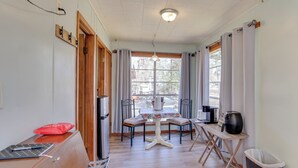 Ein gemütliches Zimmer mit einem Holztisch, einem kleinen Tisch mit floralem Tischläufer, einem Stuhl, einem Mülleimer und einem Fenster mit Vorhängen.