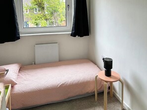 Una habitación pequeña con cama individual, ventana, radiador, una mesita con un objeto negro encima y una colcha rosa.