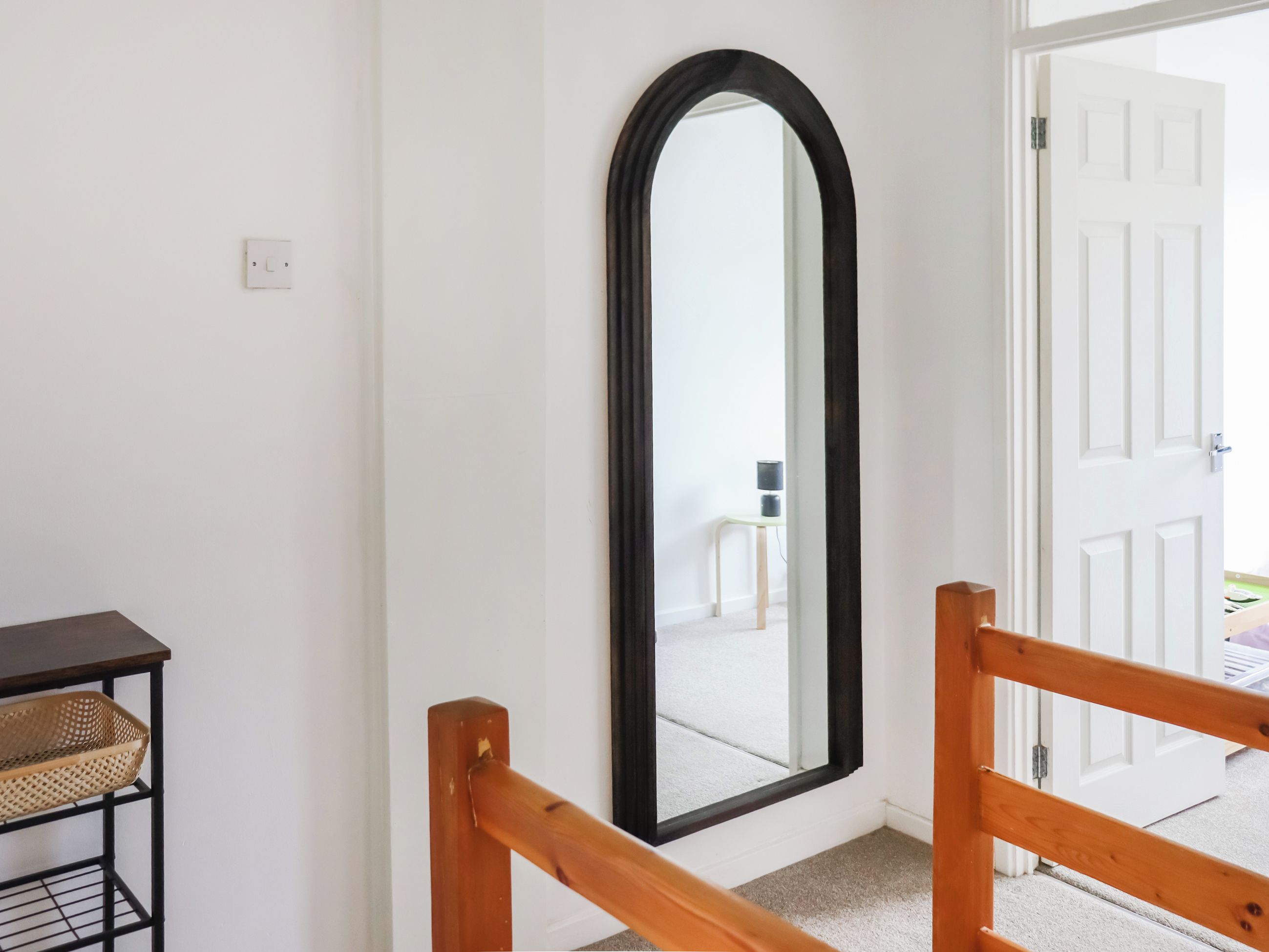A room with a wooden-framed mirror, a small table with a basket, and a door leading to another space.