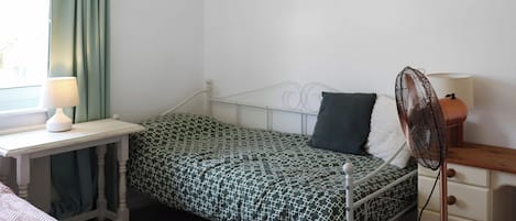 A single bed with a patterned bedspread, a bedside table with a lamp, a nightstand with a fan, and a window with curtains.