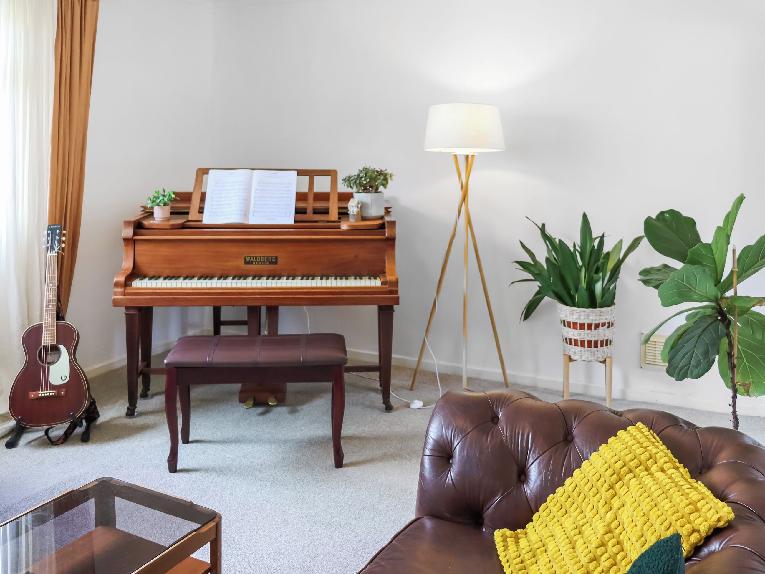 A living room with a piano, a guitar, a bench, a sofa, a floor lamp, and potted plants.