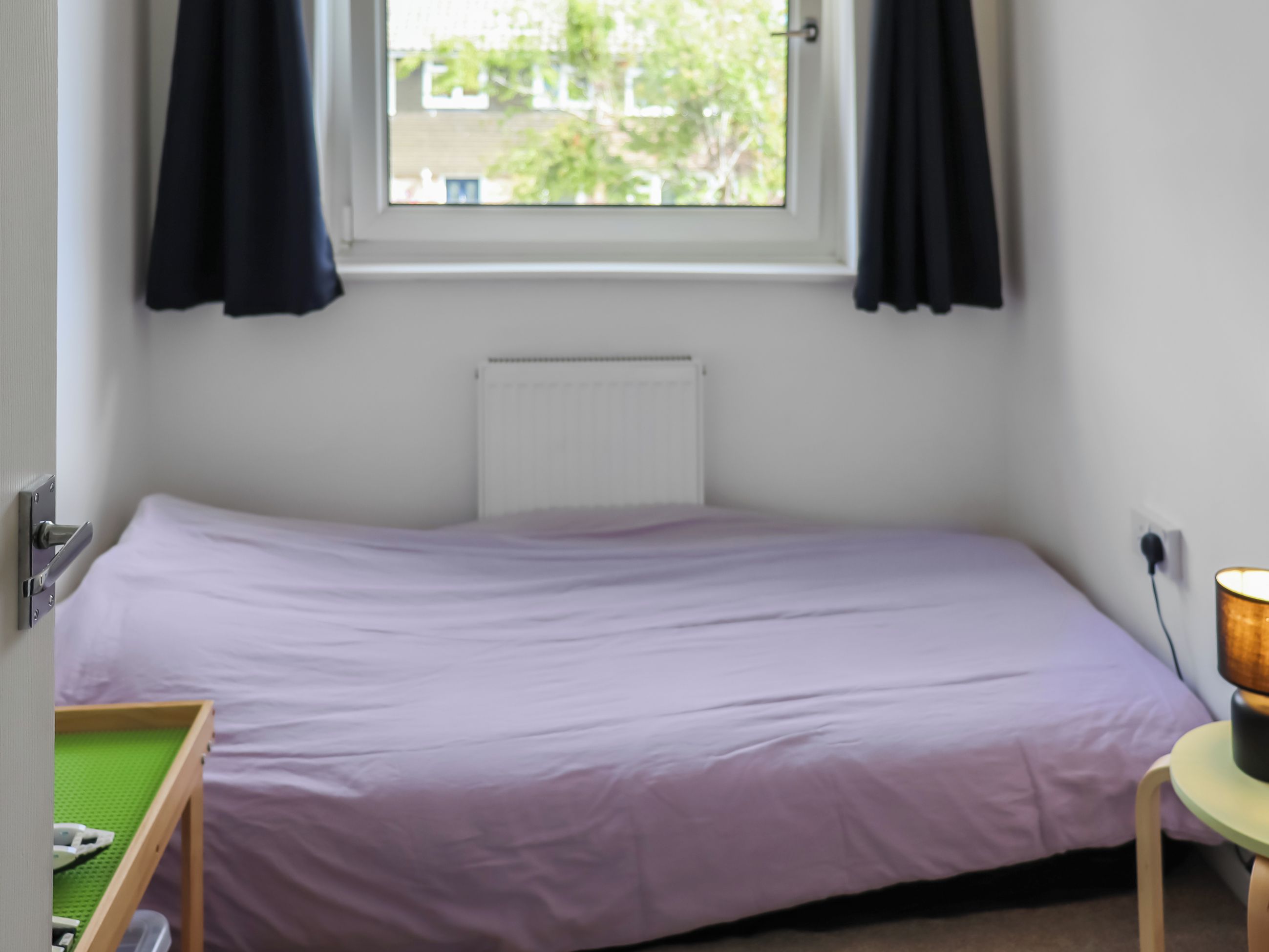 A single bed with a white headboard, a window with curtains, a bedside table with a lamp, and a small green table with a phone.