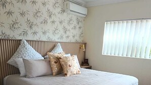 Una cama con colchón acolchado, cabecera con listones verticales, aire acondicionado fijado a la pared, ventana con persianas y papel tapiz estampado.