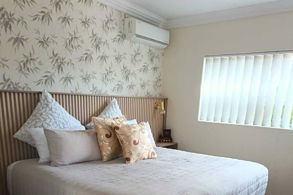 Una cama con colchón acolchado, cabecera con listones verticales, aire acondicionado fijado a la pared, ventana con persianas y papel tapiz estampado.