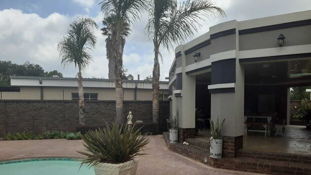 Un área de piscina con patio de ladrillo, palmeras y un edificio moderno con zona de estar cubierta al aire libre.