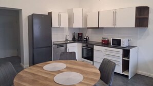 Ett kompakt kök med vita skåp, ett kylskåp, en mikrovågsugn och en kaffebryggare. Där finns också ett matbord i trä med två stolar.