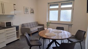 Ett kompakt kök med mikrovågsugn, en grå soffa, ett matbord med fyra stolar och ett fönster med frostat glas.