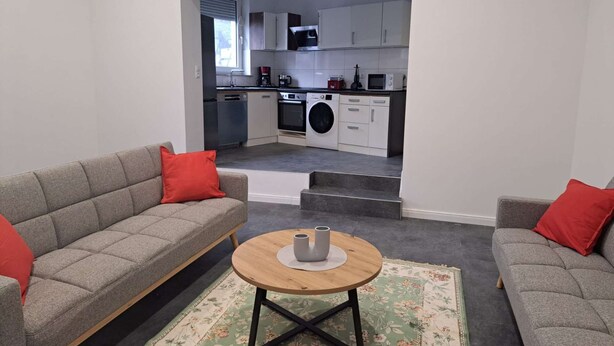 A modern living room with grey sofas and red cushions, a wooden coffee table, and a floral rug. A kitchen with white cabinets and appliances is visible through an open doorway. - Modern, spacious 3-room apartment - 85 m² (Solingen)